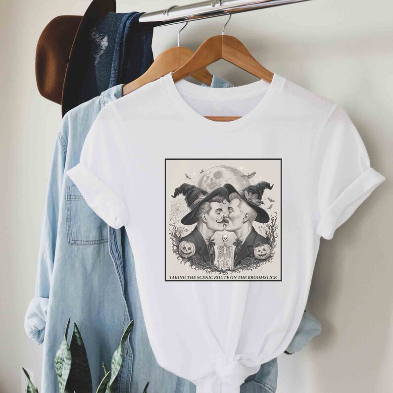 May include: White t-shirt with a black and white illustration of two men in witch hats kissing. The text "Taking the scenic route on the broomstick" is below the illustration.