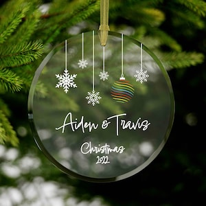 Puede incluir: Un adorno navideño de vidrio transparente con un adorno de arcoíris colgando de él. El adorno también tiene copos de nieve blancos y el texto "Aiden & Travis Navidad 2022".