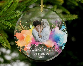 May include: A round glass Christmas ornament with a watercolor-style rainbow background. A couple is pictured in the center of the ornament, with the text "Nicole & Lana Christmas 2024" below them.