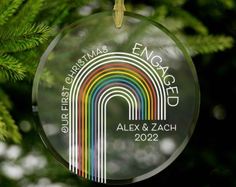 May include: A clear glass Christmas ornament with a rainbow design and the text "Our First Christmas Engaged Alex & Zach 2022".