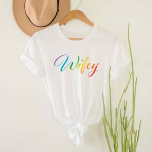 May include: White t-shirt with the word "Wifey" written in rainbow colors.