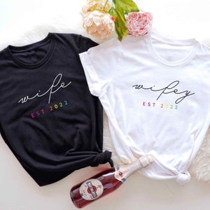 May include: Two white t-shirts with black text that says "wife" and "wifey" with rainbow colored "EST 2022" underneath.