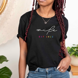 May include: Black t-shirt with the word "wife" written in white script and "EST 2022" in rainbow colors below.