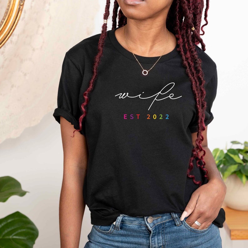 May include: Black t-shirt with the word "wife" written in white script and "EST 2022" in rainbow colors below.