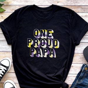 May include: Black t-shirt with the text "ONE PROUD PAPA" in a 3D style with purple, yellow, and white stripes.