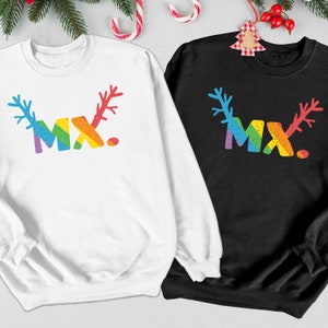 Könnte beinhalten: Zwei Weihnachtspullover, einer weiß und einer schwarz, mit einem Regenbogen-Rentier-Design und dem Text "MX."