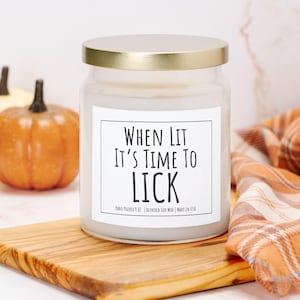 Puede incluir: Una vela en tarro de vidrio transparente con tapa dorada, con la inscripci&oacute;n "WHEN LIT IT'S TIME TO LICK". La vela est&aacute; sobre una tabla de madera, con calabazas y una bufanda a cuadros en el fondo. Hecho a mano y hecho en los EE. UU.