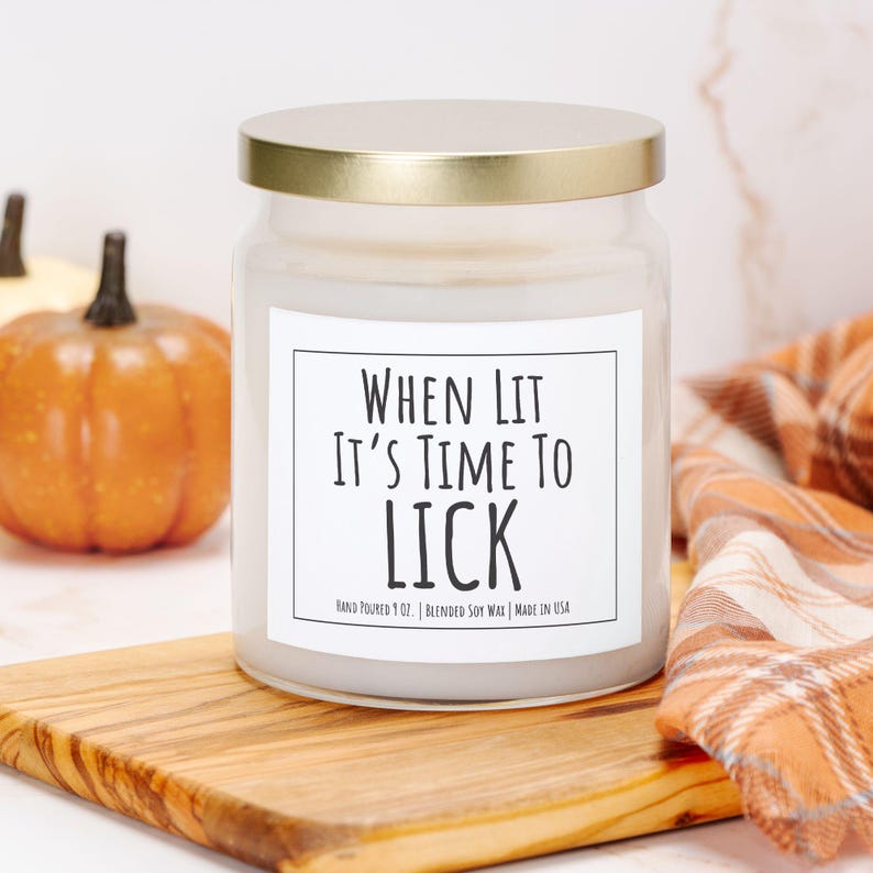 Puede incluir: Una vela en tarro de vidrio transparente con tapa dorada, con la inscripci&oacute;n "WHEN LIT IT'S TIME TO LICK". La vela est&aacute; sobre una tabla de madera, con calabazas y una bufanda a cuadros en el fondo. Hecho a mano y hecho en los EE. UU.