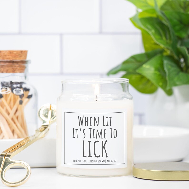 Puede incluir: Una vela de cera de soja blanca encendida en un tarro de cristal transparente con tapa dorada. La vela tiene una etiqueta que dice "WHEN LIT IT'S TIME TO LICK". Unas tijeras doradas y un tarro de cerillas est&aacute;n en el fondo.