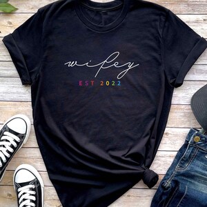 May include: Black t-shirt with the word "wifey" written in white cursive script and "EST 2022" written in rainbow colors below.