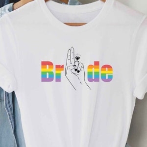 Puede incluir: Camiseta blanca con la palabra "Bride" en colores arcoíris. La letra "i" se reemplaza por una mano con un anillo de diamantes en el dedo.