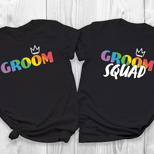 May include: Two black t-shirts. One says "GROOM" in rainbow colours, the other "GROOM SQUAD" in white with rainbow accents. Both have a small white crown above the text. The shirts are made of a soft material.