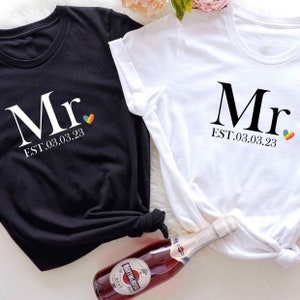 Op de afbeelding: Twee zwarte en witte T-shirts met het woord "Mr." en de datum "EST. 03.03.23" erop gedrukt. Een regenboog hartje staat naast de datum op elk shirt.