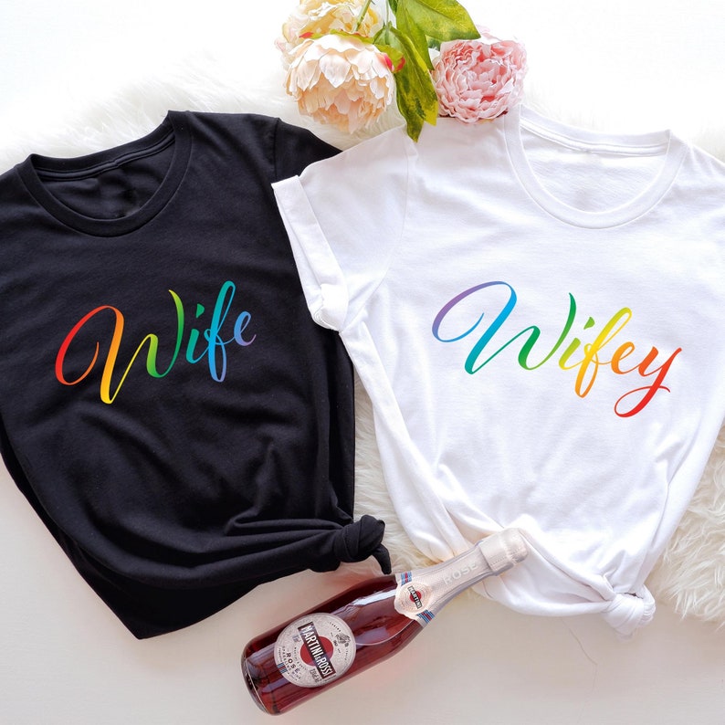 May include: Two t-shirts with the words "Wife" and "Wifey" written in rainbow colors. The shirts are black and white and are lying on a white surface.