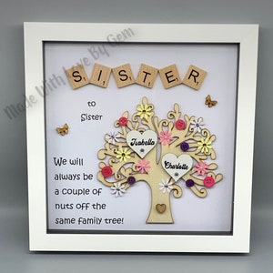 May include: A white framed picture with a wooden tree design. The tree has pink, yellow, and purple flowers and buttons. The words "Sister" and "to Sister" are spelled out with wooden letter tiles. The text "We will always be a couple of nuts off the same family tree!" is written below the tree. The names "Isabella" and "Charlotte" are written on hearts on the tree.