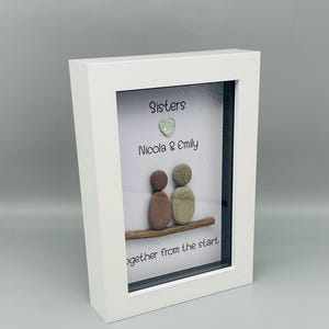 Puede incluir: Una obra de arte enmarcada en blanco con la palabra "Sisters" y los nombres "Nicola & Emily". Dos figuras de guijarros est&aacute;n sentadas en una rama de madera, con un coraz&oacute;n brillante encima de los nombres. El texto "together from the start" est&aacute; abajo.