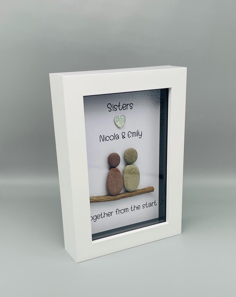 Puede incluir: Una obra de arte enmarcada en blanco con la palabra "Sisters" y los nombres "Nicola & Emily". Dos figuras de guijarros est&aacute;n sentadas en una rama de madera, con un coraz&oacute;n brillante encima de los nombres. El texto "together from the start" est&aacute; abajo.