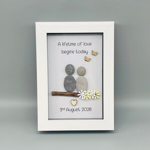 May include: A framed artwork with a white border. The artwork features two pebble figures, a wooden branch, and decorative elements including butterflies, flowers, and a heart. The text reads "A lifetime of love begins today" and "3rd August 2026".