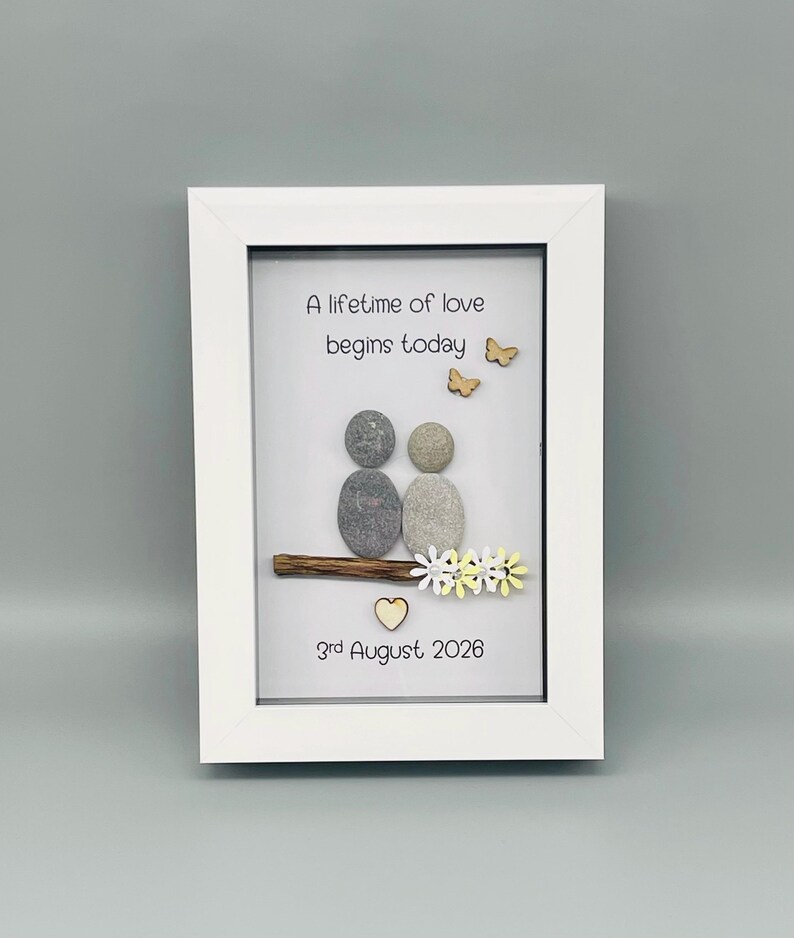 May include: A framed artwork with a white border. The artwork features two pebble figures, a wooden branch, and decorative elements including butterflies, flowers, and a heart. The text reads "A lifetime of love begins today" and "3rd August 2026".