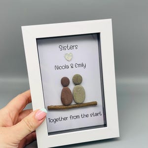 Puede incluir: Una obra de arte enmarcada en blanco. Presenta dos figuras de guijarros sobre una rama de madera, con las palabras "Sisters", "Nicola & Emily" y "Together from the start". Un peque&ntilde;o coraz&oacute;n brillante est&aacute; encima de los nombres.