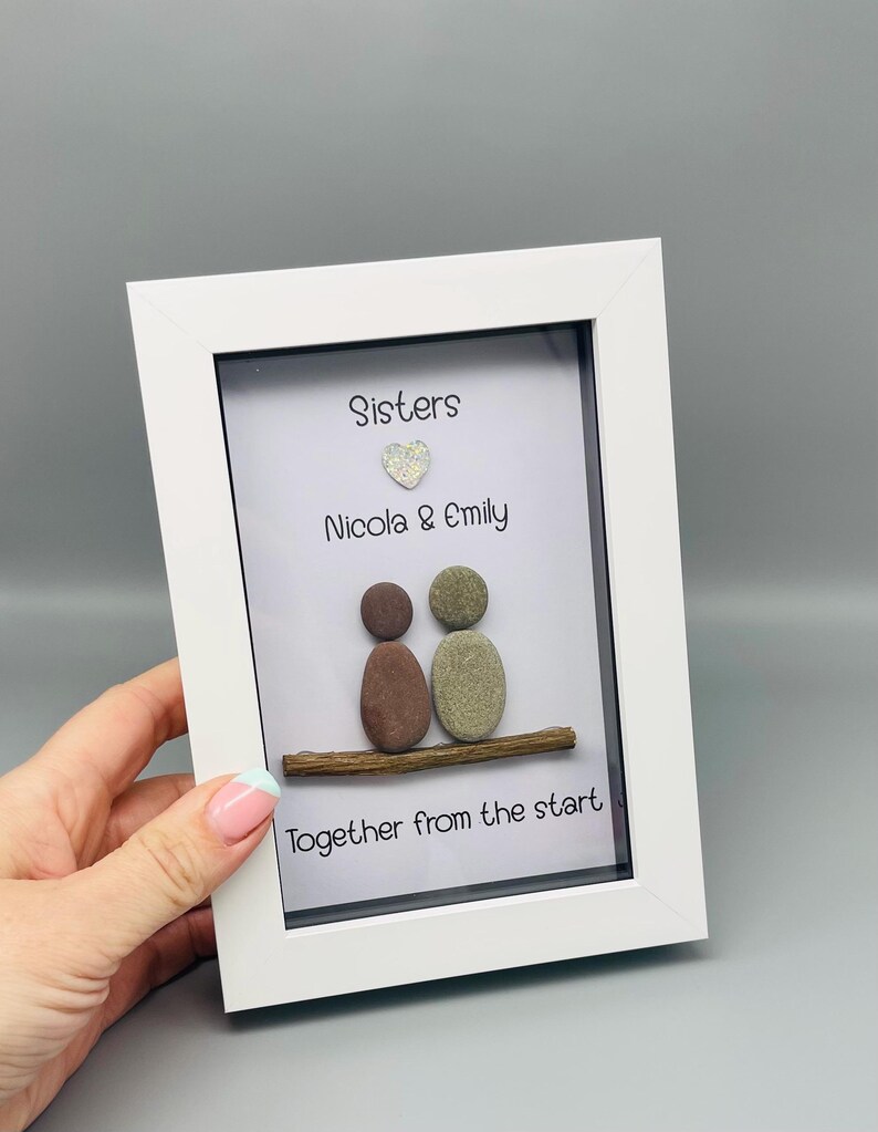 Puede incluir: Una obra de arte enmarcada en blanco. Presenta dos figuras de guijarros sobre una rama de madera, con las palabras "Sisters", "Nicola & Emily" y "Together from the start". Un peque&ntilde;o coraz&oacute;n brillante est&aacute; encima de los nombres.