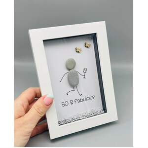May include: White framed art piece with a pebble figure holding a champagne glass. The art includes the text "50 & fabulous" and two small wooden butterflies. The bottom of the art is decorated with clear crystals.