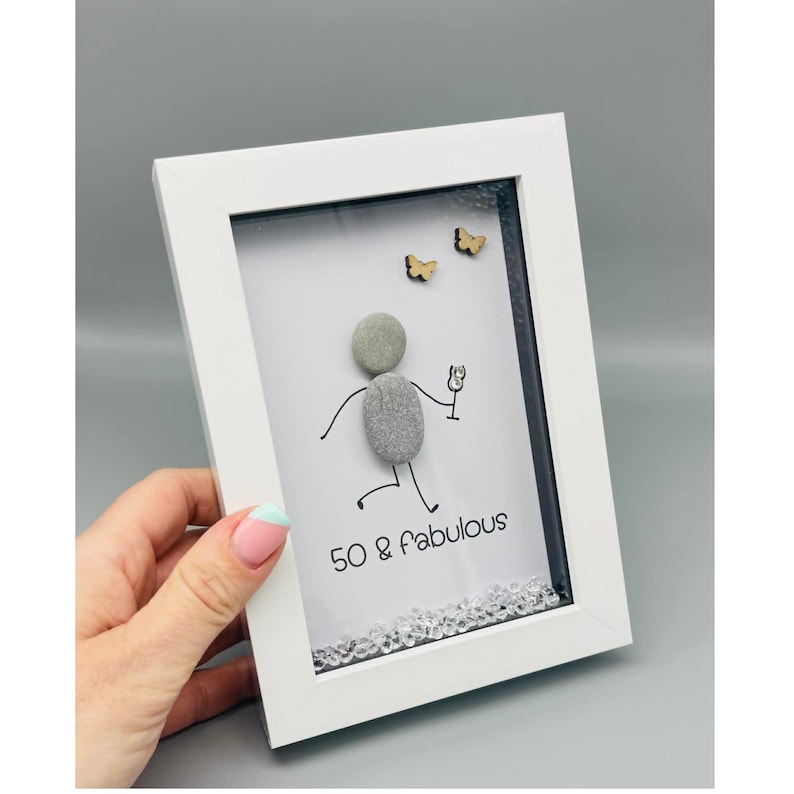 May include: White framed art piece with a pebble figure holding a champagne glass. The art includes the text "50 & fabulous" and two small wooden butterflies. The bottom of the art is decorated with clear crystals.