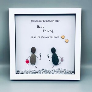 May include: White framed artwork with a hand-painted message that reads "Sometimes being with your best friend is all the therapy you need." The artwork features two stick figures made of stones holding glasses of wine. The stick figures are standing on a bed of rhinestones.