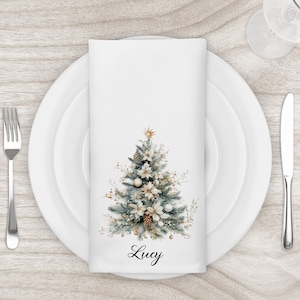 May include: A white cloth napkin with a watercolor Christmas tree design, set on a white plate with silverware. The tree features gold and white ornaments, pinecones, and a star. The name "Lucy" is written in script at the bottom.