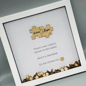 May include: A white picture frame with a white background and a quote about love. The quote reads "There's over 7 billion people on this planet And in a heartbeat I'd still choose you". The frame also includes two wooden puzzle pieces with the names "Emma" and "Logan" written on them.