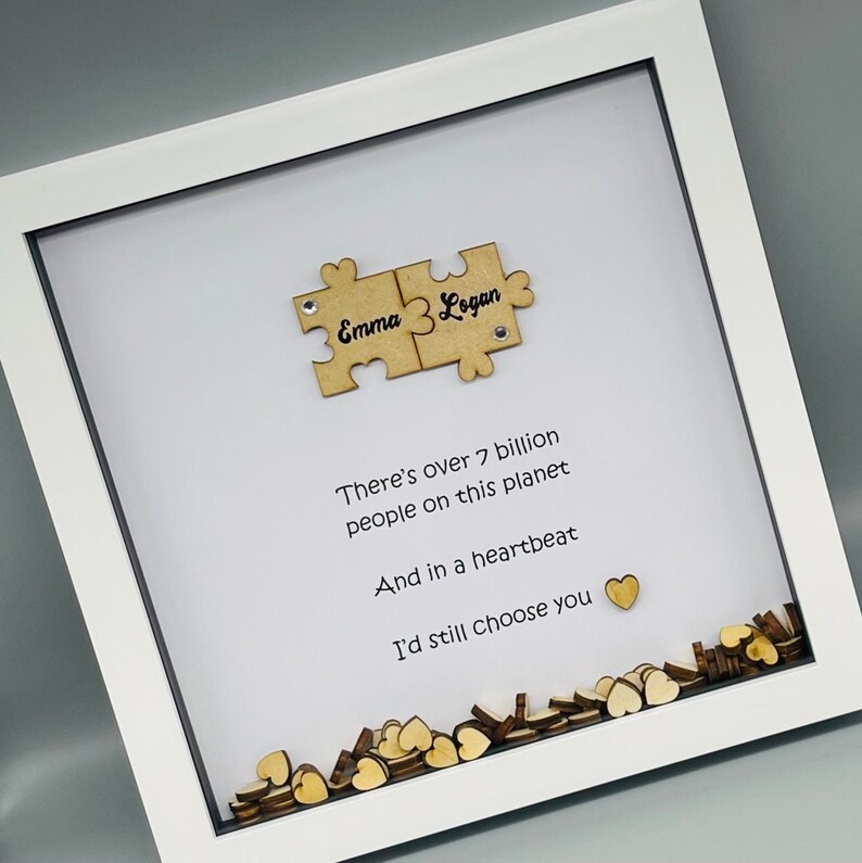 May include: A white picture frame with a white background and a quote about love. The quote reads "There's over 7 billion people on this planet And in a heartbeat I'd still choose you". The frame also includes two wooden puzzle pieces with the names "Emma" and "Logan" written on them.