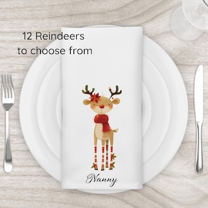 May include: A white napkin featuring a cartoon reindeer with a red scarf and striped legs. The napkin is on a white plate, with a fork and knife. Text on the napkin reads "12 Reindeers to choose from" and "Nanny".