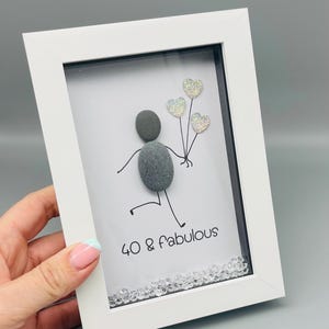 Op de afbeelding: Een ingelijst kunstwerk met een steenkunstfiguur die hartvormige ballonnen vasthoudt. De figuur is gemaakt van grijze kiezels. De tekst "40 & fabulous" staat eronder. De lijst is wit en de achtergrond is wit.
