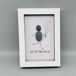 Op de afbeelding: Een witgelijst kiezelkunstwerk. Het toont een stokfiguur gemaakt van grijze kiezels die glitter hartballonnen vasthoudt. De tekst "40 & fabulous" staat eronder. Het frame is wit.
