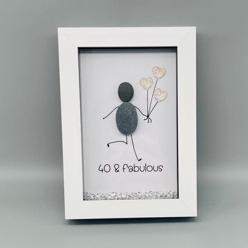 Op de afbeelding: Een witgelijst kiezelkunstwerk. Het toont een stokfiguur gemaakt van grijze kiezels die glitter hartballonnen vasthoudt. De tekst "40 & fabulous" staat eronder. Het frame is wit.