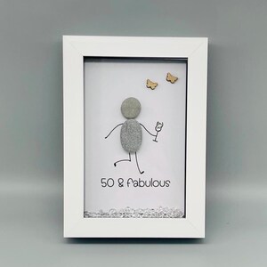 May include: A framed artwork with a white border. The artwork features a stick figure made of gray stones holding a wine glass, with the text "50 & fabulous". Two wooden butterflies are above the figure.