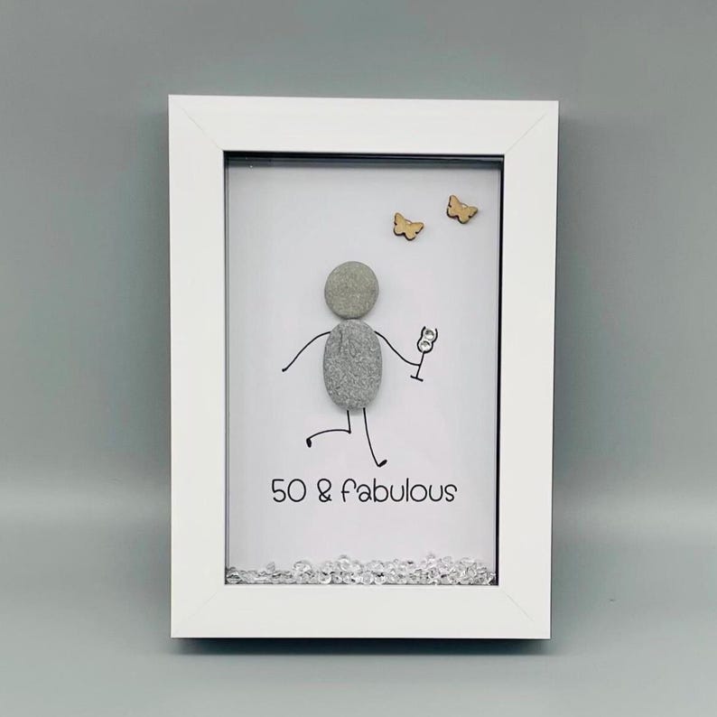 May include: A framed artwork with a white border. The artwork features a stick figure made of gray stones holding a wine glass, with the text "50 & fabulous". Two wooden butterflies are above the figure.