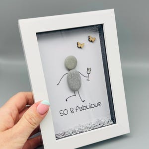 May include: A framed art piece with a white border. The artwork features a pebble person holding a glass, with the text "50 & Fabulous" below. Two small wooden butterflies and decorative crystals complete the design.