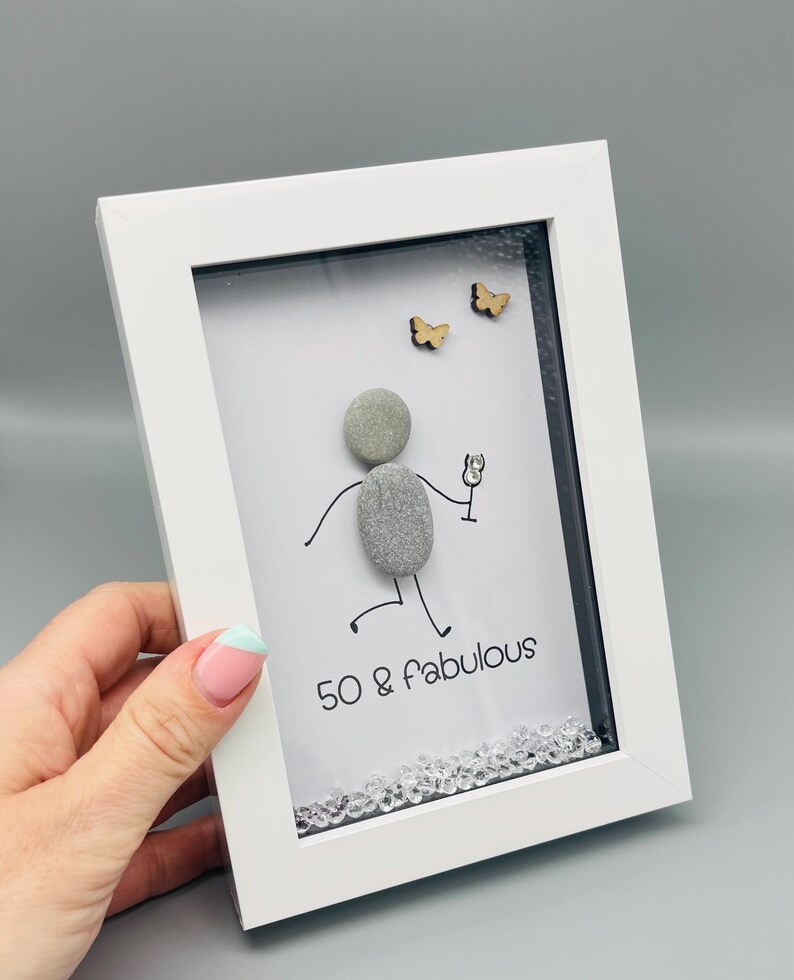 May include: A framed art piece with a white border. The artwork features a pebble person holding a glass, with the text "50 & Fabulous" below. Two small wooden butterflies and decorative crystals complete the design.