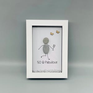 May include: A framed pebble art piece with a stick figure holding a wine glass, celebrating a 50th birthday. The text "50 & fabulous" is below the figure. Two small wooden butterflies are above the figure. The frame is white.