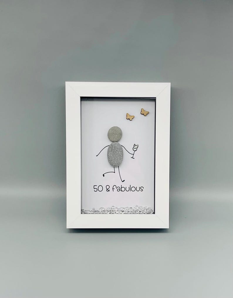 May include: A framed pebble art piece with a stick figure holding a wine glass, celebrating a 50th birthday. The text "50 & fabulous" is below the figure. Two small wooden butterflies are above the figure. The frame is white.