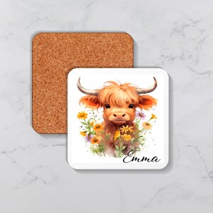 Op de afbeelding: Een witte keramische onderzetter met een aquarel-illustratie van een Highland-koe met bruine vacht en een wit gezicht. De koe is omringd door gele en paarse bloemen. De tekst "Emma" staat in zwart onder de koe.