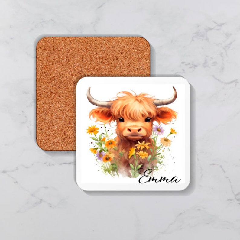 Op de afbeelding: Een witte keramische onderzetter met een aquarel-illustratie van een Highland-koe met bruine vacht en een wit gezicht. De koe is omringd door gele en paarse bloemen. De tekst "Emma" staat in zwart onder de koe.
