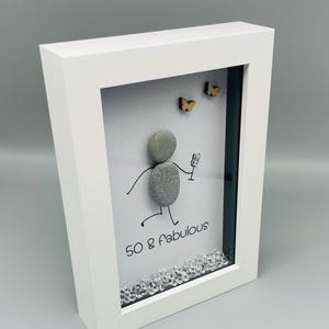 May include: White framed art piece with a pebble figure holding a glass, with the text "50 & fabulous". Two small wooden butterflies are above the figure. Clear crystals are at the bottom of the frame.