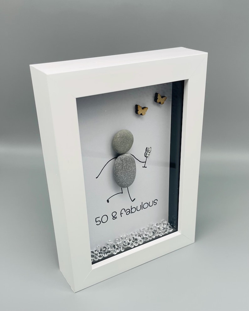 May include: White framed art piece with a pebble figure holding a glass, with the text "50 & fabulous". Two small wooden butterflies are above the figure. Clear crystals are at the bottom of the frame.