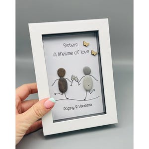May include: White framed artwork with the text "Sisters, A lifetime of love." The art features pebble figures holding glasses, with the names "Poppy & Vanessa" below. Two small wooden butterflies are also included.