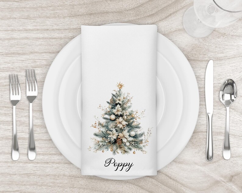May include: A white cloth napkin with a watercolor Christmas tree design, featuring gold and white ornaments, pinecones, and a star. The word "Poppy" is written in script at the bottom. The napkin is set on a white plate with silverware.
