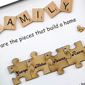 Peut inclure: Un cadre blanc avec un fond blanc et une citation qui dit "FAMILY are the pieces that build a home". La citation est entour&eacute;e de lettres de jeu en bois qui &eacute;pellent "FAMILY" et un petit papillon en bois. Sous la citation, il y a quatre pi&egrave;ces de puzzle en bois qui s'embo&icirc;tent. Chaque pi&egrave;ce de puzzle a un nom &eacute;crit dessus : George, Abbie, Florence et Rosie.
