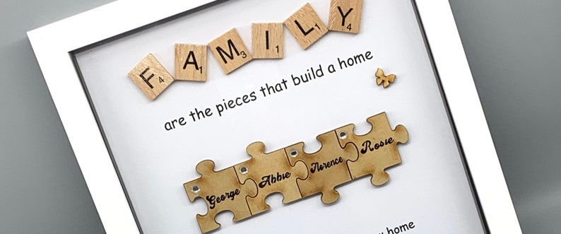 Peut inclure: Un cadre blanc avec un fond blanc et une citation qui dit "FAMILY are the pieces that build a home". La citation est entour&eacute;e de lettres de jeu en bois qui &eacute;pellent "FAMILY" et un petit papillon en bois. Sous la citation, il y a quatre pi&egrave;ces de puzzle en bois qui s'embo&icirc;tent. Chaque pi&egrave;ce de puzzle a un nom &eacute;crit dessus : George, Abbie, Florence et Rosie.