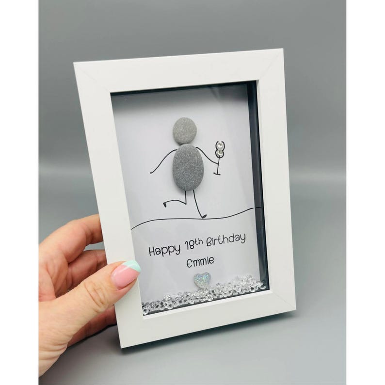 Peut inclure: Cadre blanc avec une &oelig;uvre d'art en galets repr&eacute;sentant une personne tenant un verre. Le texte dit "Happy 18th Birthday Emmie" avec un c&oelig;ur argent&eacute; et des cristaux. Le cadre est tenu par une main.